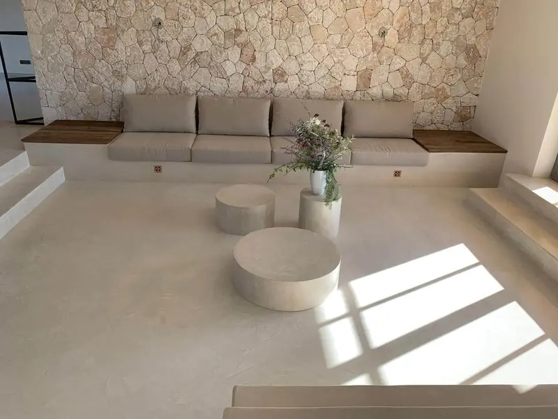 Salón con sofá de obra, mesas cilíndricas y suelo de microcemento bajo una pared de piedra